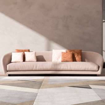 Curved sofa upholstered in light beige fabric with coral and rust throw pillows on a dark wood base, against a rose-beige wall.