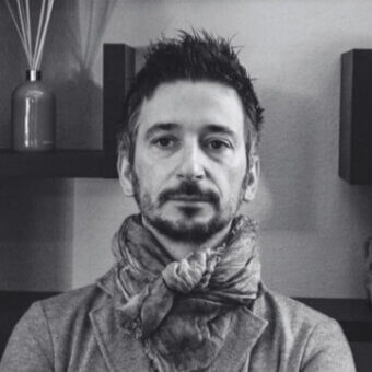 Black and white portrait of Alessandro Corro wearing a gray sweater and textured scarf, with a diffuser and boxes on a shelf behind him.