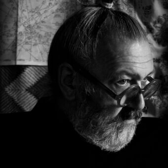 Black and white portrait of Costantino Morosin in profile, wearing glasses and a gray beard, with a blurred vintage map background.