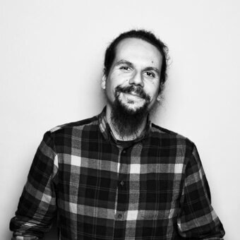 Alberto Ghirardello portrait, black and white photograph, man with long dark hair, beard, and plaid shirt, neutral expression, soft lighting.