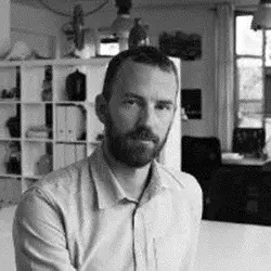 Black and white portrait of Alberto Manzoni seated at a white table, with a shelving unit and window in the background.