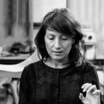 Beatriz Sempere, woman with dark hair, gesturing while speaking, wearing a dark textured top, blurred interior background, soft lighting.
