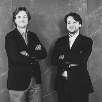 Black and white portrait of two men with crossed arms, positioned before gray-veined marble panels, wearing suits and casual attire.