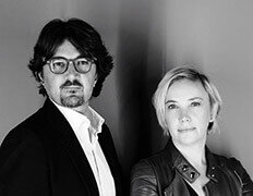 Black and white portrait of two designers, man in suit, woman in leather jacket, against dark background, studio lighting.