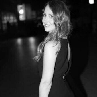 Black and white portrait of woman in sleeveless dress, silver bracelet, looking over shoulder with blurred nighttime background.