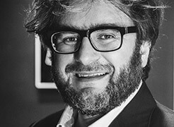 Daniele Basso portrait, black and white photograph, man with beard wearing glasses and dark suit jacket, blurred background.