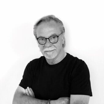 Black and white portrait of Danilo de Rossi, a man with gray hair, glasses, and a black t-shirt, arms crossed against a white wall.