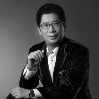 Man in black velvet suit jacket and glasses, three-quarter portrait, dark watch visible, gray gradient background, composed expression.