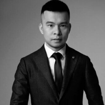 Man in dark tweed suit, white shirt, black tie, neutral gray background, formal portrait, serious expression, notched lapel, single-button closure.