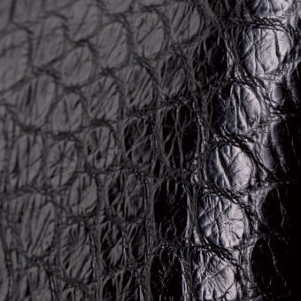 Black alligator skin texture with dense, raised ridges and irregular depressions, displaying a subtle sheen and monochromatic tone.