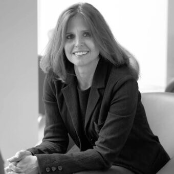 Mariela Gensler portrait, woman with wavy light brown hair, wearing a black top and gray tailored blazer, soft diffused lighting.