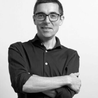 Black and white portrait of a man with short hair, glasses, and a dark button-down shirt, arms crossed, with “patrick jouin id” text.