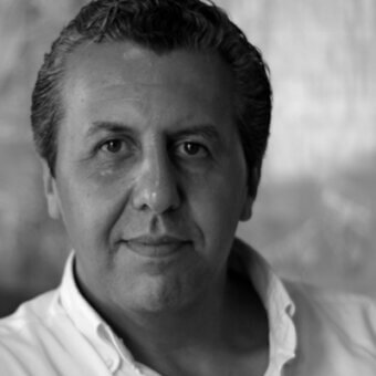 Black and white portrait of Riccardo Giovanetti, a man with short dark hair, wearing a white button-down shirt, against a blurred interior background.