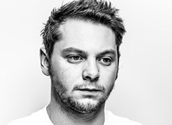 Black and white portrait of Romain Le Mahieu, a man with short dark hair, trimmed beard, and neutral expression against a light gray backdrop.