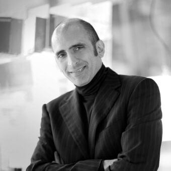 Man in dark pinstripe suit, black turtleneck, smiling; blurred architectural background, soft studio lighting, professional portrait.
