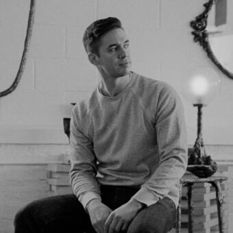 Man seated on books, wearing gray sweatshirt and jeans, against brick wall with rope and lamp on stacked books.