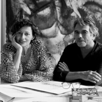 Black and white portrait of two artists in studio, woman in patterned blouse, man in sweater, abstract art background.