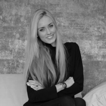 Woman with blonde hair seated on white cushion against gray mottled wall, wearing black turtleneck and bracelets.