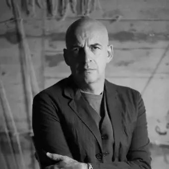 Bald man in gray blazer against weathered wood wall, arms crossed, wearing a wristwatch, dramatic lighting, Antonio Marras portrait.