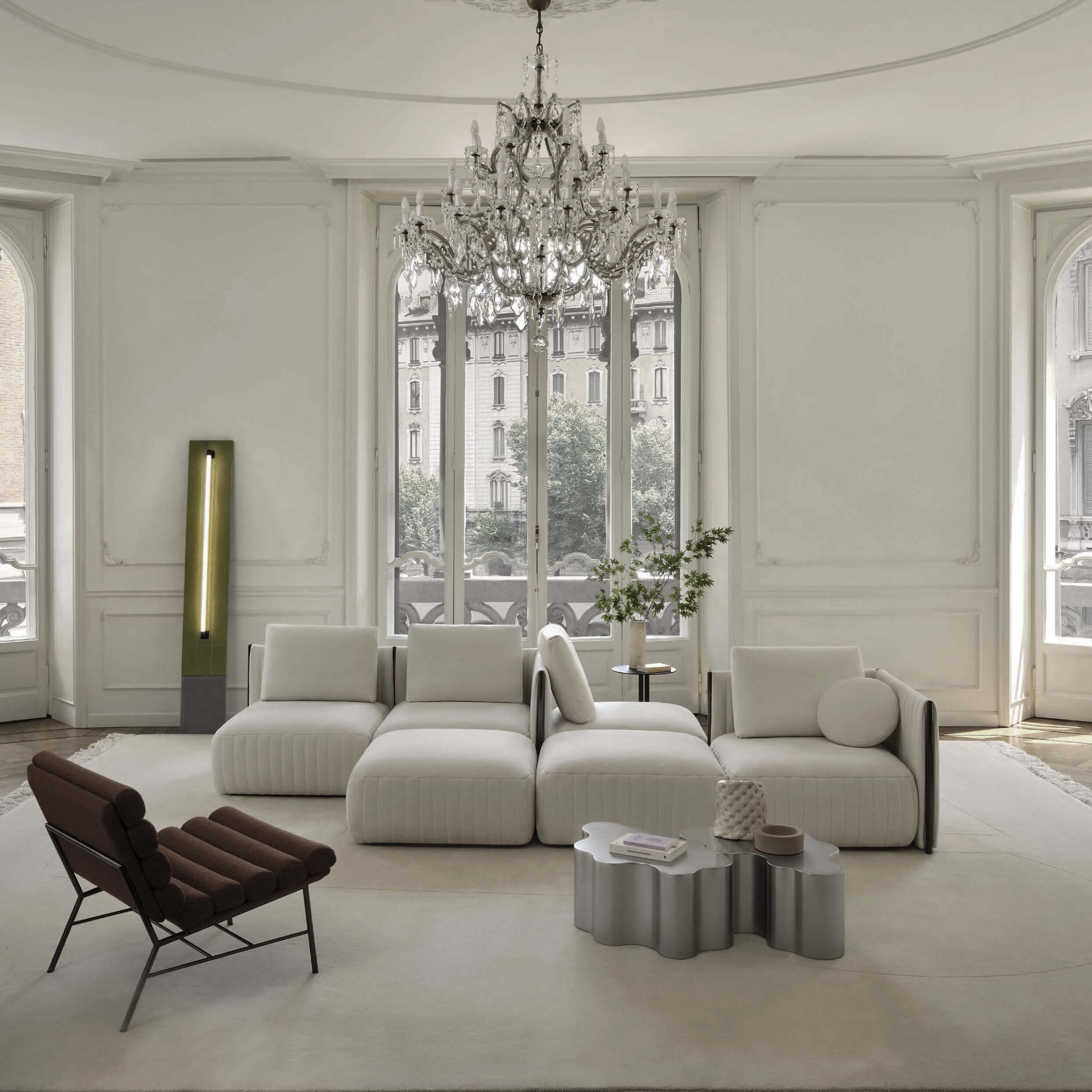 Cream modular sofa with wide armrests in a living room featuring a crystal chandelier, leather armchair, silver coffee table, and floor lamp.