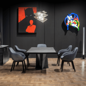 Dining table with dark gray matte top and black pedestal, surrounded by six gray velvet chairs in a room with Batman-themed artwork and crystal chandelier.