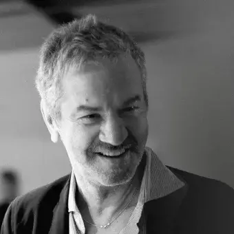 Black and white portrait of a man with gray hair, a beard, and a striped shirt under a dark blazer, wearing a silver chain.