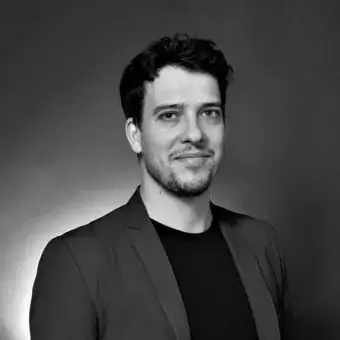 Black and white portrait of Nicola Pavan against gray gradient, wearing black t-shirt and dark gray blazer, facing left with short beard.
