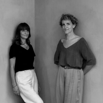 Black and white photograph of two women standing against a pale textured wall, wearing wide-leg trousers and casual tops.