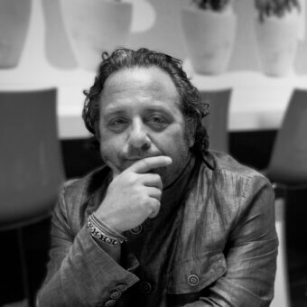 Man in olive blazer seated in modern interior, hand on chin, wearing beaded bracelet, blurred background with chairs and planters.