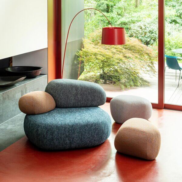 Blue armchair and beige stools with rounded forms sit on a polished floor, alongside a red arched floor lamp in a modern interior with garden view.