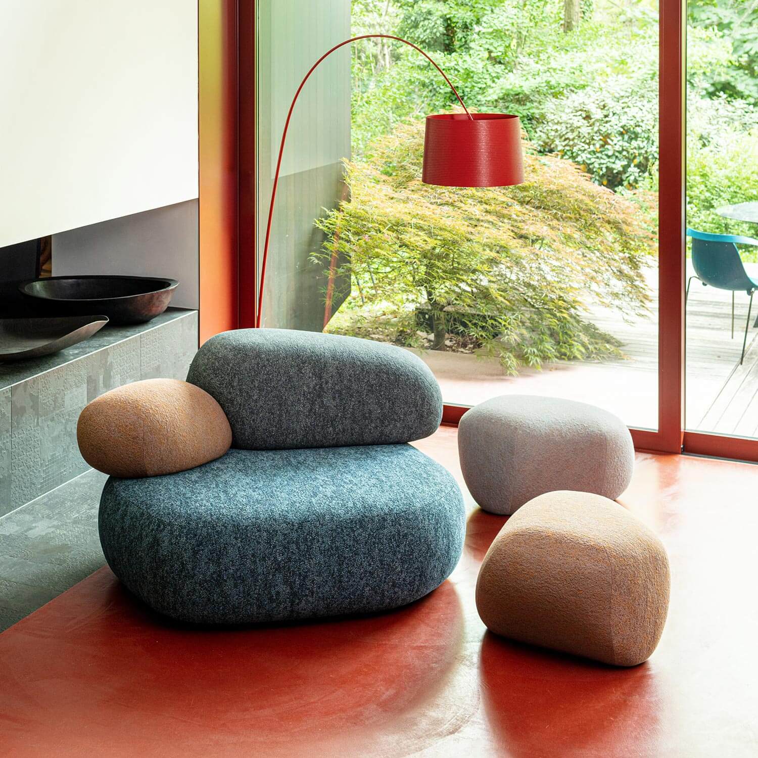 Blue armchair and beige stools with rounded forms sit on a polished floor, alongside a red arched floor lamp in a modern interior with garden view.