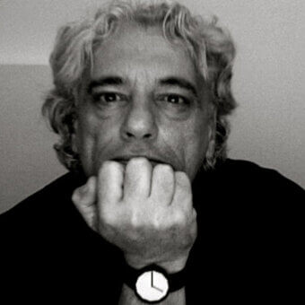 Portrait of Bruno Rainaldi, a man with gray curly hair, wearing a wristwatch with a white face and black strap, against a light gray backdrop.