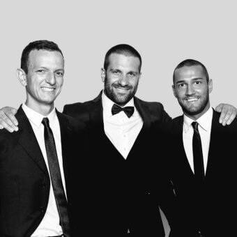 Black and white portrait of three formally dressed men in suits, centered composition, light gray backdrop, studio lighting, subtle text detail.