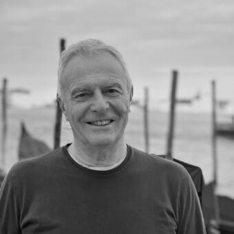 Luciano Marson portrait, grayscale, middle-aged man with gray hair wearing a dark sweater, blurred waterfront background.