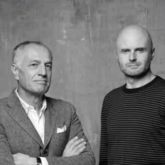 Black and white portrait of two men against a gray textured wall, one in a tweed blazer, the other in a striped shirt.