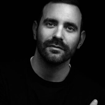 Matteo Zorzenoni portrait, black and white photograph, man with dark hair and beard, wearing black shirt, hand gesture.