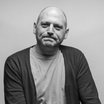Black and white portrait of a man with shaved head, salt-and-pepper beard, gray t-shirt, and dark gray cardigan, neutral background.