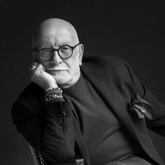 Man in charcoal blazer seated in chair, wearing glasses, ring, bracelets, against dark backdrop, portrait with focused lighting.
