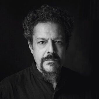 Black and white portrait of Paolo Darrigo, a man with curly dark hair, gray beard, wearing a dark collared shirt, direct gaze.