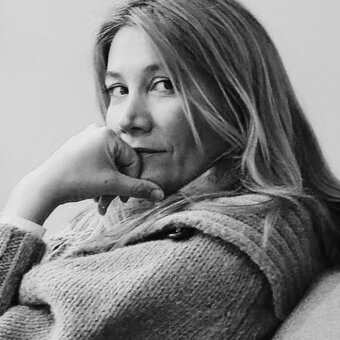 Raffaella Mangiarotti portrait, black and white, woman with long hair wearing a textured knit sweater, contemplative expression, gray background.