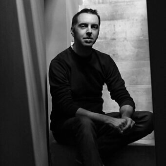 Black and white portrait of Stefano Gaggero, seated in a dark sweater, against a textured concrete wall with strong vertical shadows.