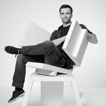 Modern chair light wood geometric design, studio portrait of man tim kerp seated casually in black t-shirt and jeans against white backdrop.