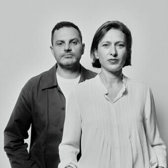 Portrait of two people, a man in dark blue shirt and a woman in light shirt, against a light gray backdrop, facing forward with neutral expressions.