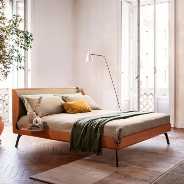 Bolzan Letti gabri bed, beige fabric headboard, rust frame, brass legs, gray linen bedding, green throw, arched floor lamp, hardwood floor.