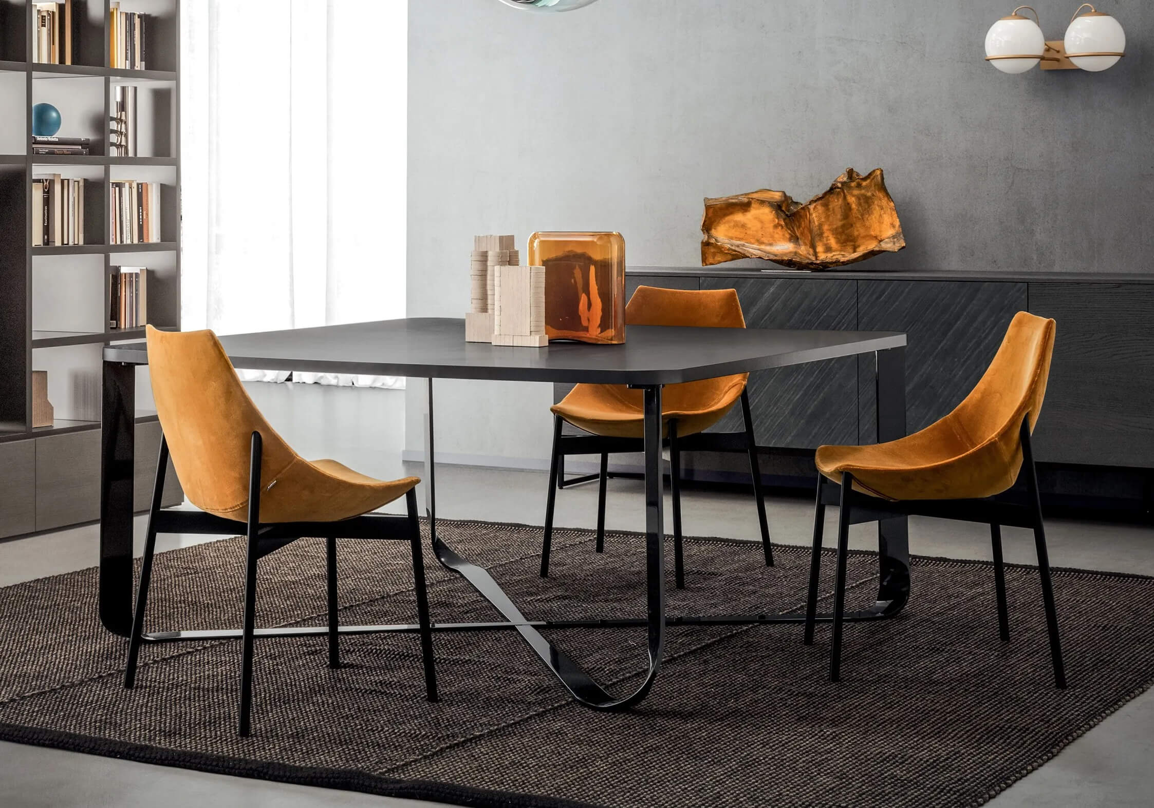 Dining table with dark gray matte top and intersecting metal base, surrounded by brown leather chairs on a dark brown rug.