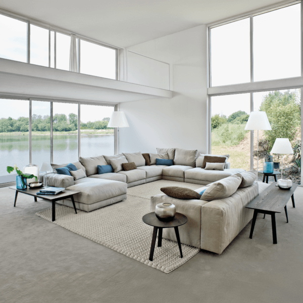 Beige modular sectional sofa with teal pillows in a modern living room featuring water views, wood tables, and a cream rug.