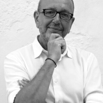 Black and white portrait of Emilio Nanni, a man with short gray beard wearing glasses and white shirt, hand raised in contemplation.