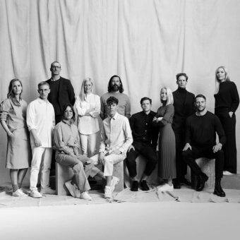 Black and white group portrait of thirteen people in neutral clothing against a draped fabric backdrop, staggered formation, soft lighting.