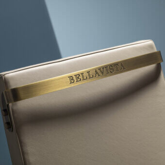 Chair handle, beige leather texture, with central brushed brass band displaying “BELLAVISTA” text, curved form, studio lighting.
