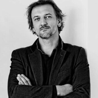 Portrait of Xavier Lust, a man with graying dark hair, wearing a dark sweater and tweed blazer, against a light gray backdrop.